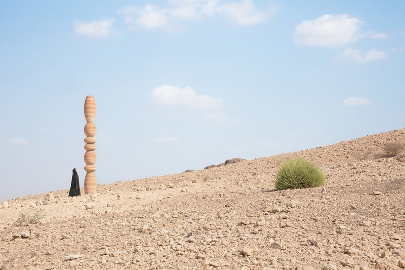 Single laminated wood column standing in rocky terrain with a figure in black robes