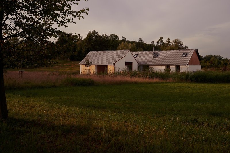 Linked gabled buildings with tile roofs and timber cladding viewed across a meadow in evening light