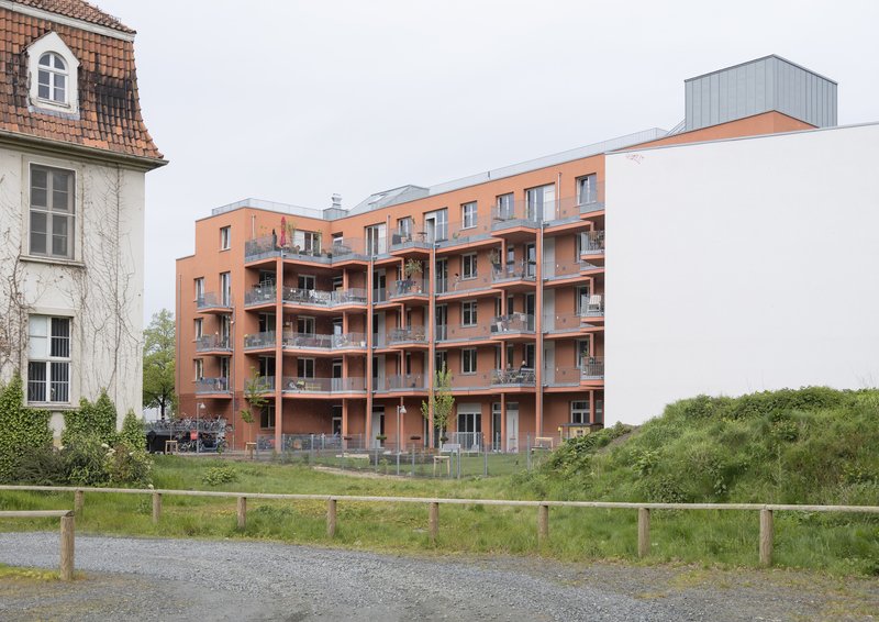 Terracotta brick facade with recessed balconies flanked by older buildings and a grassy field under overcast skies