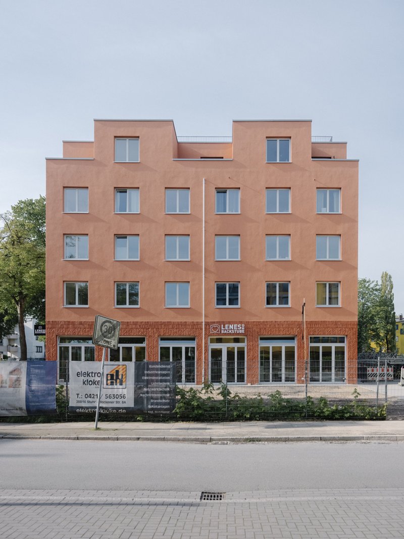 Street facade with terracotta plaster, punched windows, stepped parapet, and ground-floor retail beneath an overcast sky