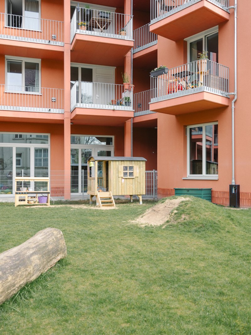 Coral-colored facade with recessed balconies above a shared lawn with a small wooden playhouse