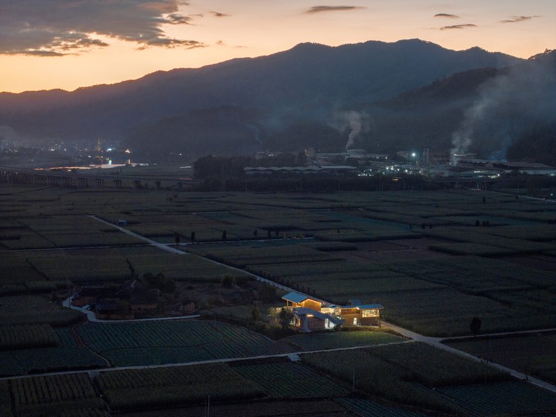 Illuminated residential complex in farmland at dusk with industrial buildings and smoke plumes visible
