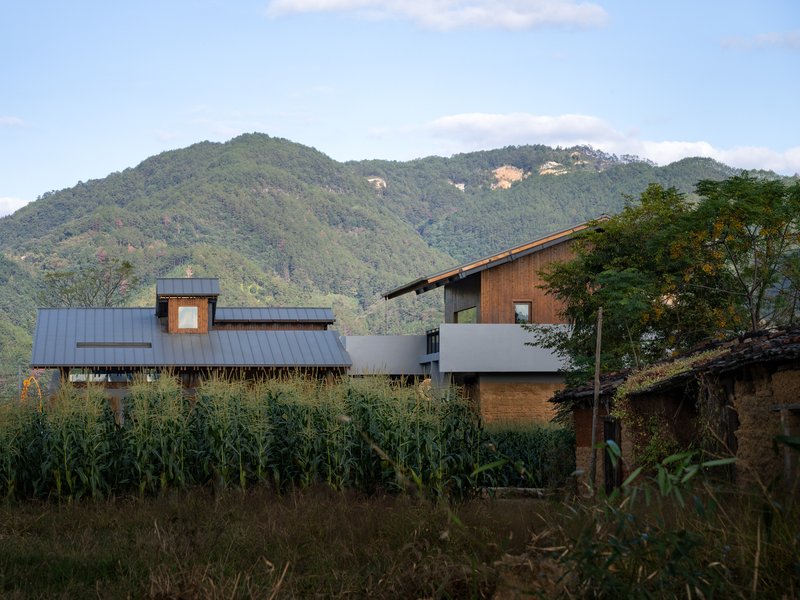 Mixed-material building complex with timber and metal cladding set among cornfields with forested mountains beyond