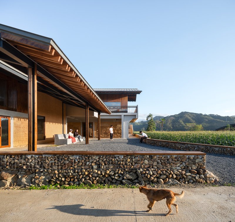 Gabion retaining wall with stone fill supporting a gravel terrace where a dog walks below the eaves