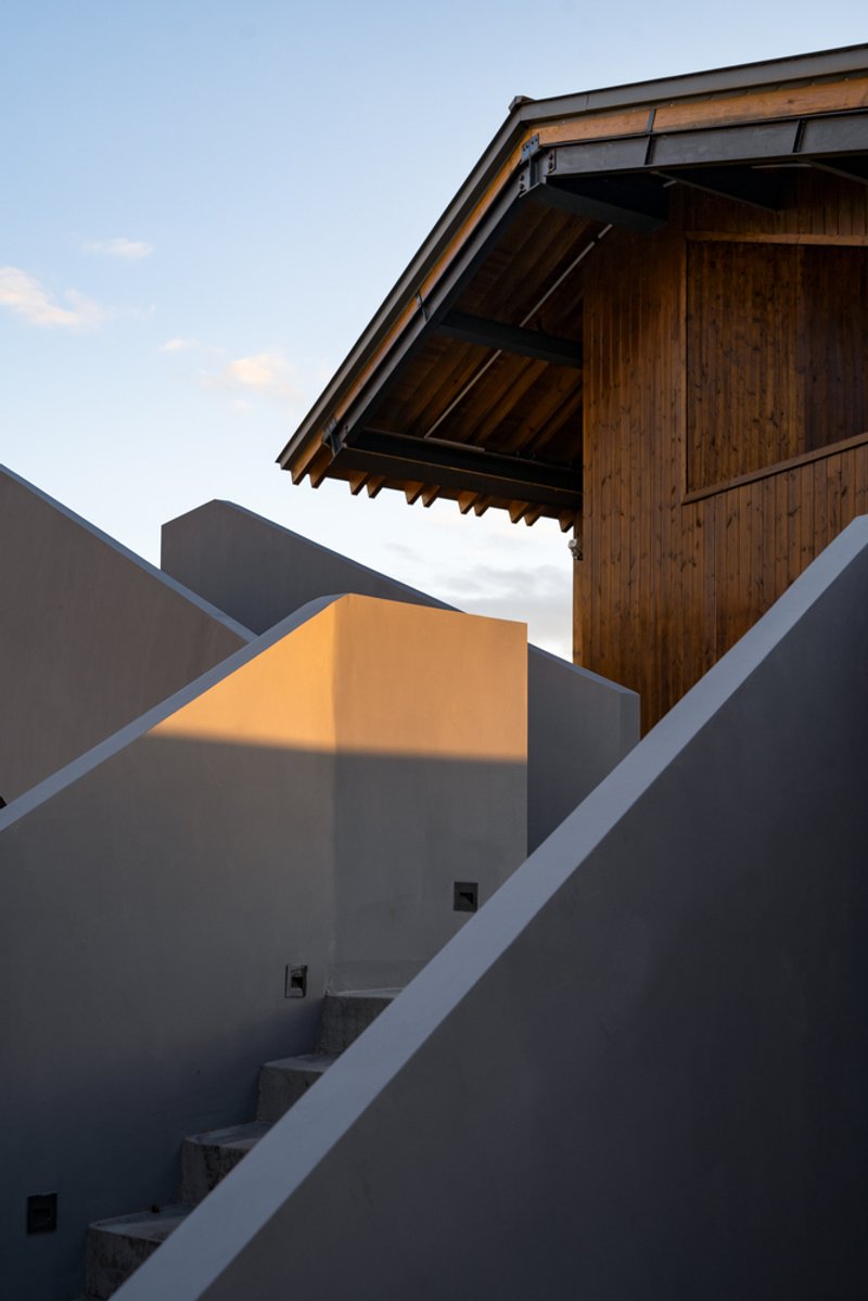 Concrete stair ascending through angular rendered walls toward a timber-clad pavilion in evening light
