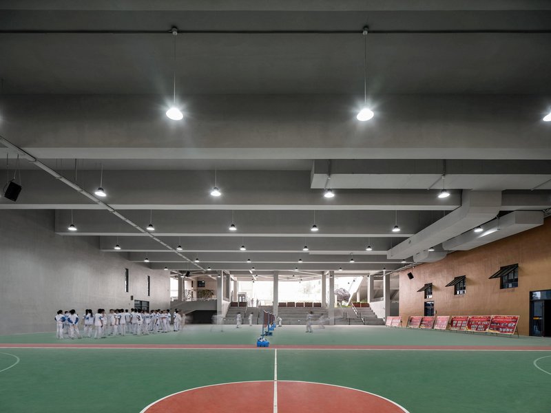 Indoor sports court with exposed concrete ceiling beams and pendant lights where students gather in martial arts uniforms