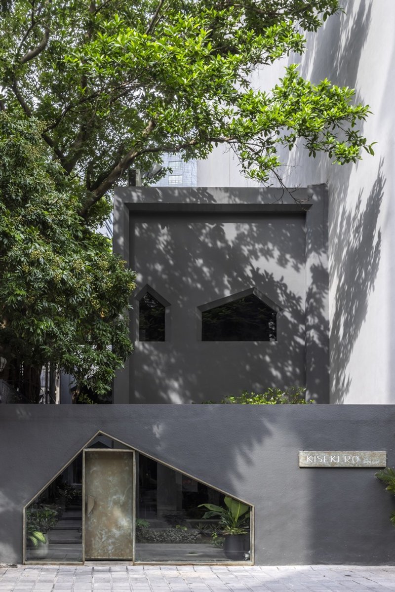 Street entrance with peaked metal gate beneath a cantilevered volume casting tree shadows