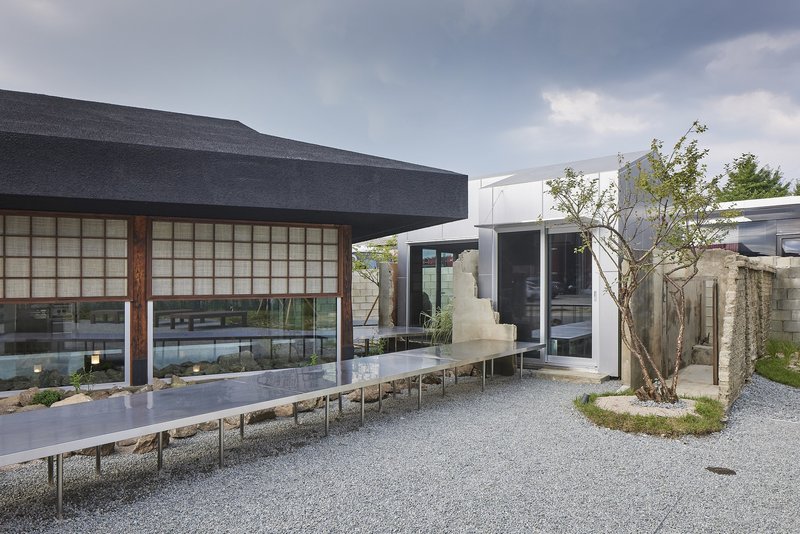 Gravel courtyard showing the pavilion with gridded facade, elevated steel walkway, and planted tree at dusk