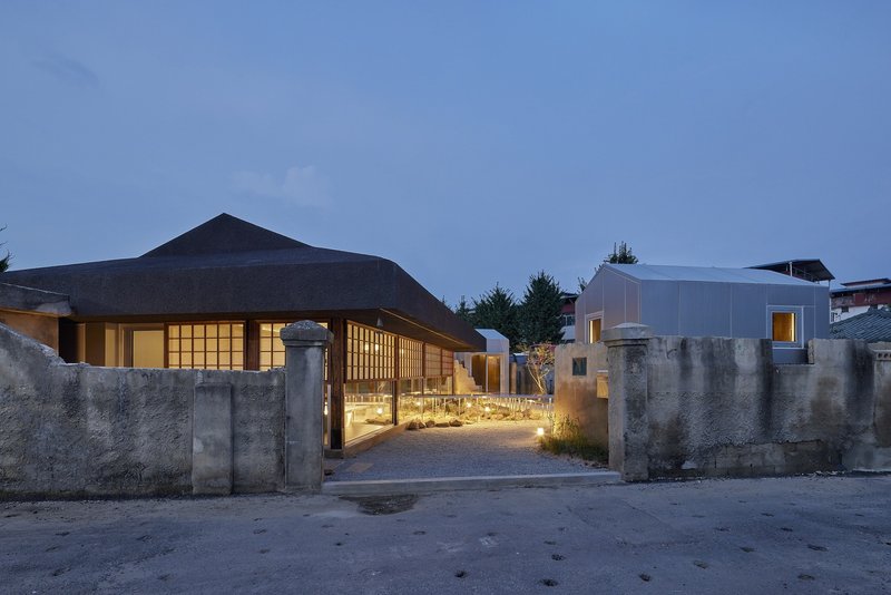 Entry courtyard with gravel path, concrete walls, and illuminated timber-framed pavilions at dusk