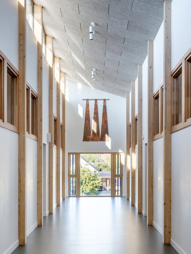 Double-height corridor with timber-framed walls and acoustic ceiling panels leading to glazed doors and courtyard beyond