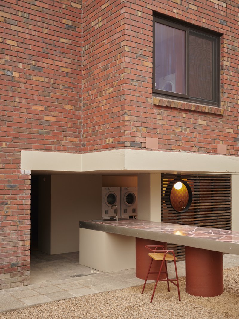 Ground-floor communal laundry niche with white bench counter and red cylindrical base beneath variegated brick wall