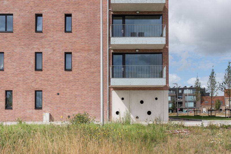 White concrete base with circular openings beneath stacked balconies and red brick facade in summer grasses