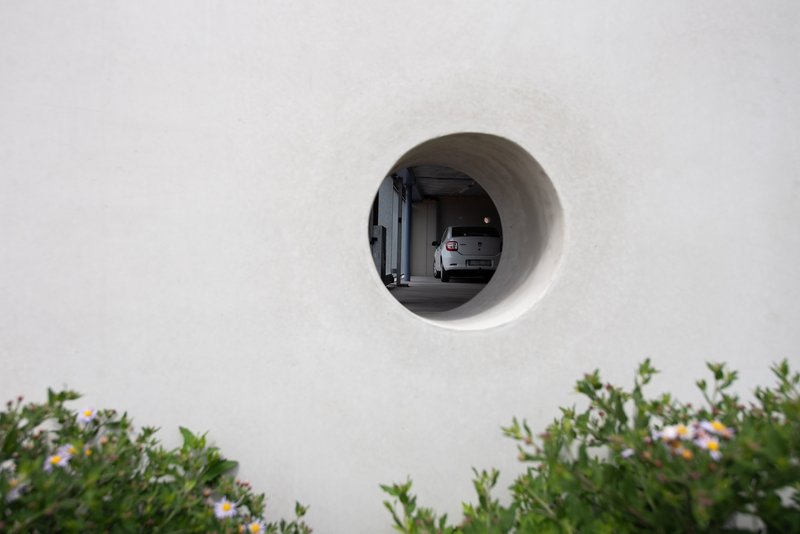 Circular opening in white wall revealing parked car beyond, with flowering shrubs below