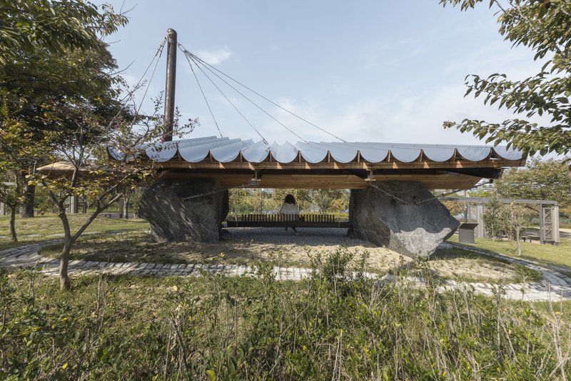 Open pavilion with undulating roof supported by large boulders and timber beams in a park setting