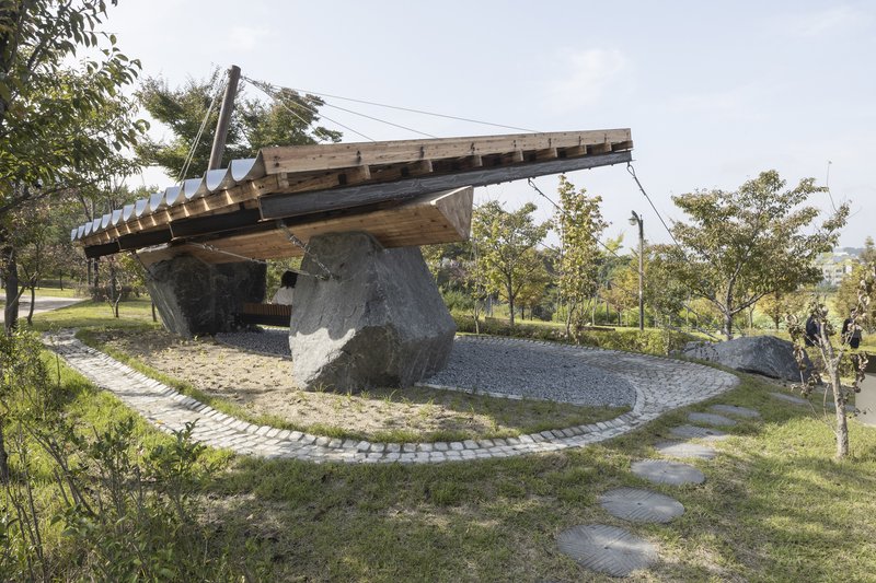 Side view of the angled roof pavilion with cable supports among scattered trees and gravel pathways