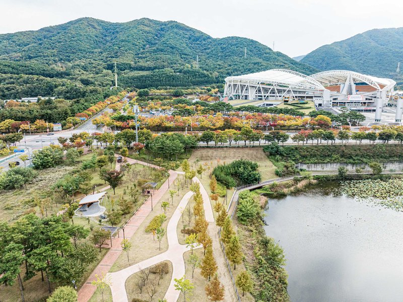 Elevated view of the riverside park with curving paths and a stadium complex beyond forested hills