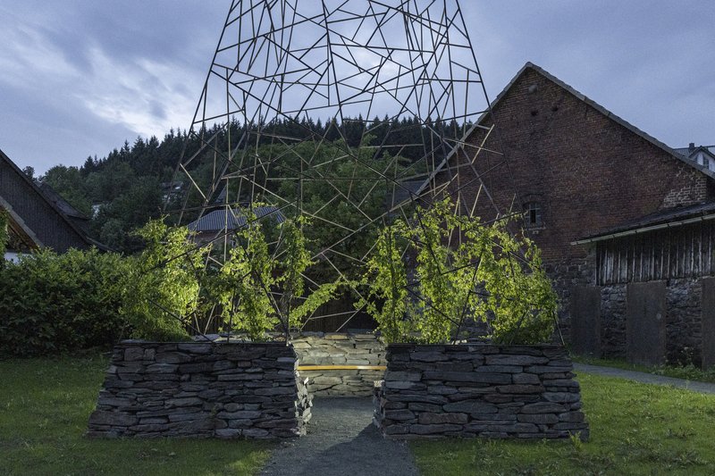 Geometric steel framework sheltering young trees planted in the layered stone enclosure at dusk