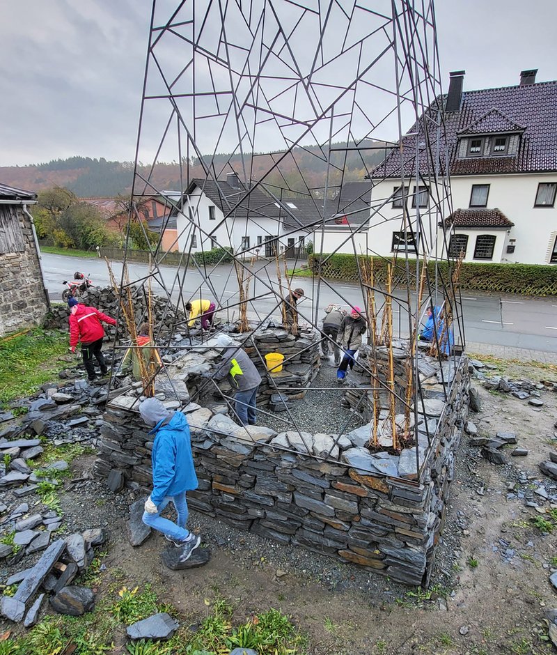 Circular dry-stone wall base with triangulated metal frame tower under construction, workers in rain gear