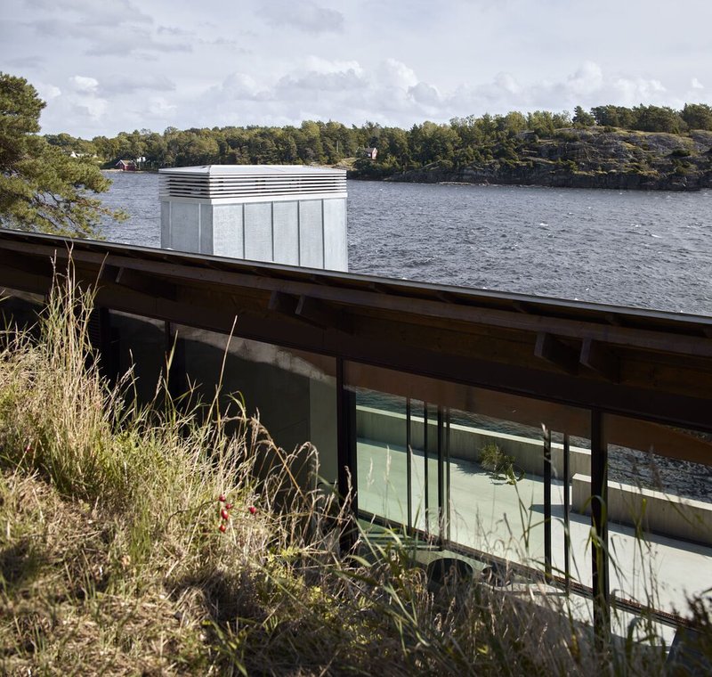 Low-angled view of the glass-walled volume with flat roof nestled into rocky terrain beside the water