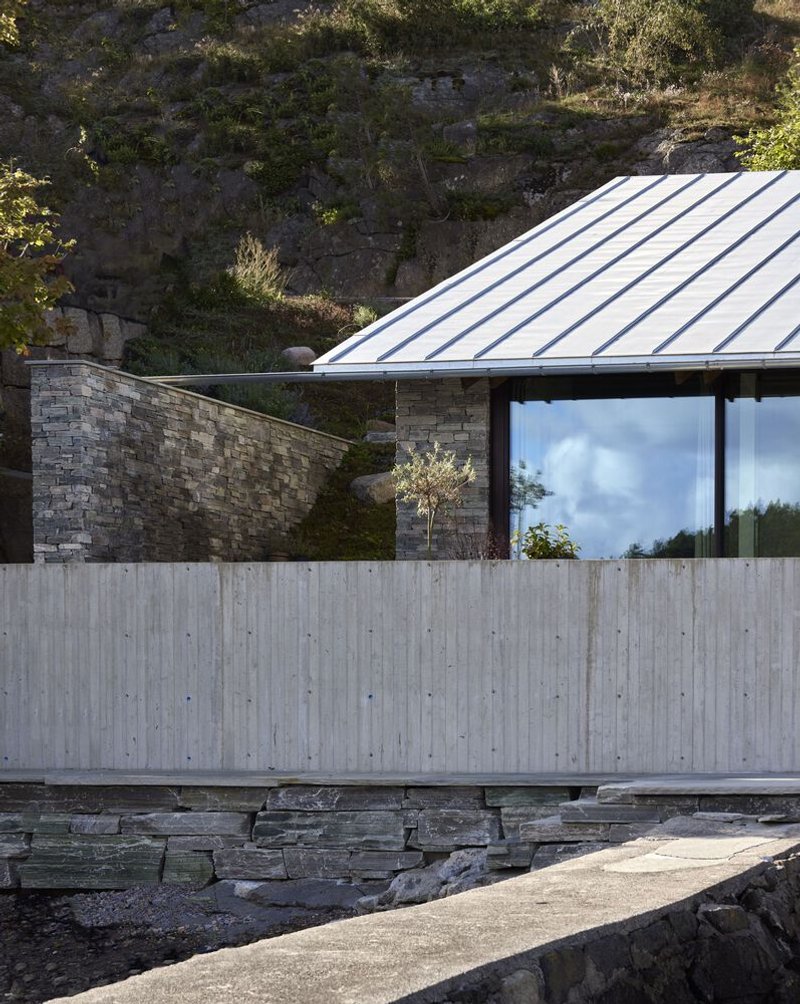 Board-formed concrete wall and stacked stone base beneath a sloped metal roof against hillside rock