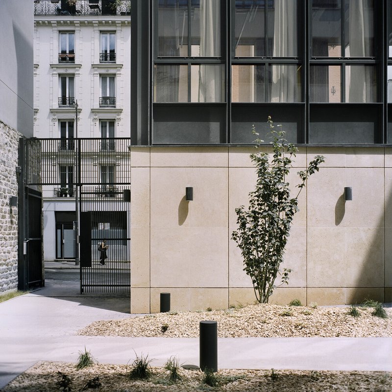 Street view of the beige stone facade with planted gravel bed and a young tree beside the entry passage