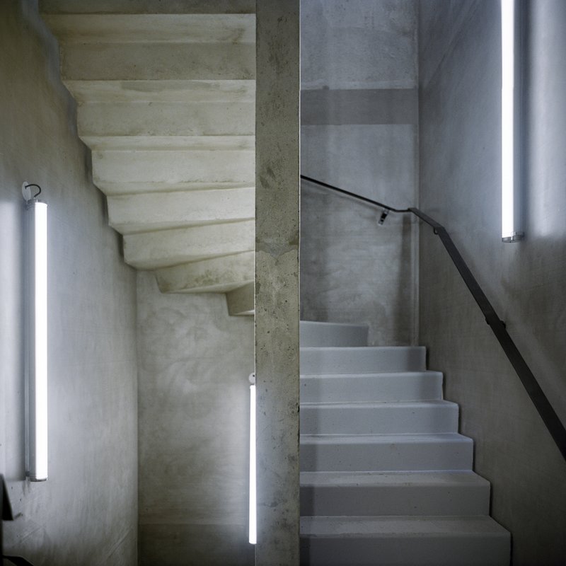 Concrete stairwell with vertical tube lighting and black metal handrail along gray plastered walls