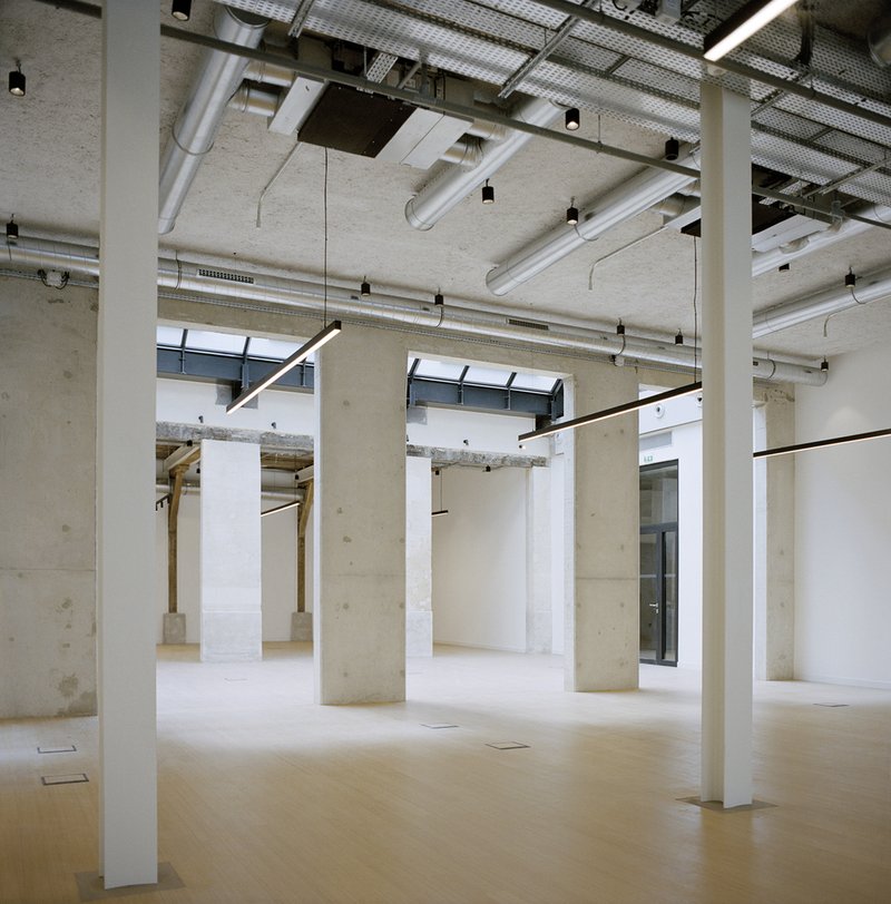 Open interior space with exposed concrete columns and ceiling ducts beneath a perforated metal screen skylight