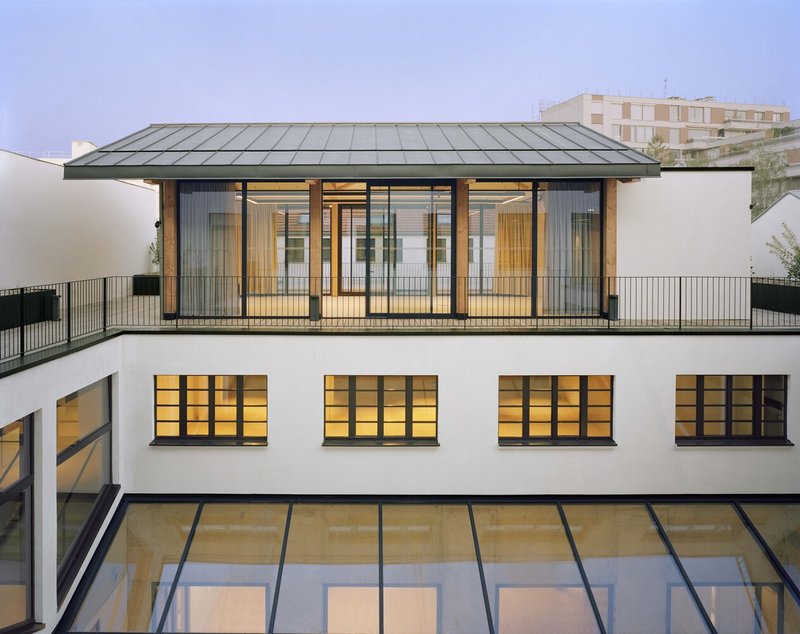 Courtyard view showing white rendered walls, glazed roof, metal-framed rooftop pavilion with timber screens at dusk