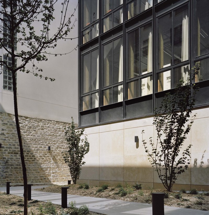 Gridded glass facade rising above stone base wall with young trees and climbing vines in the forecourt