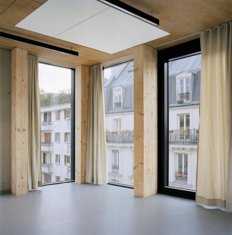 Interior corner with timber columns and exposed ceiling beams framing tall windows overlooking neighboring facades