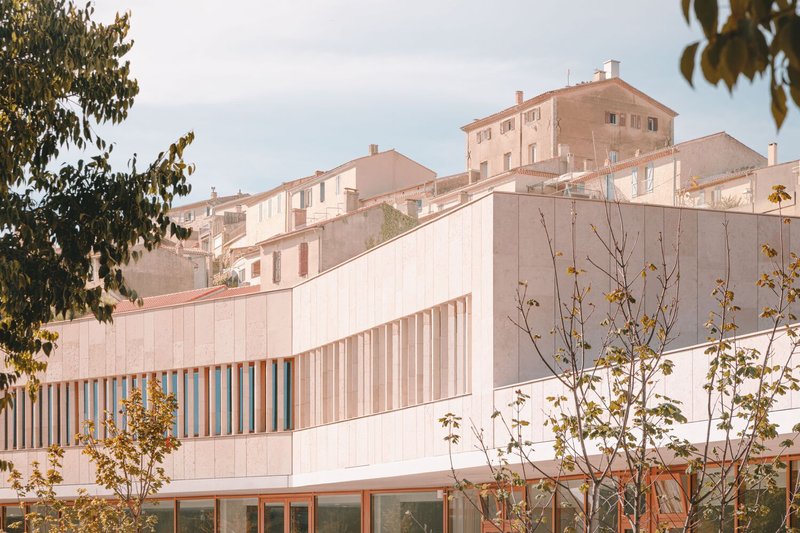 Pale ribbed facade with vertical sunshading louvers set against hillside village houses at dusk