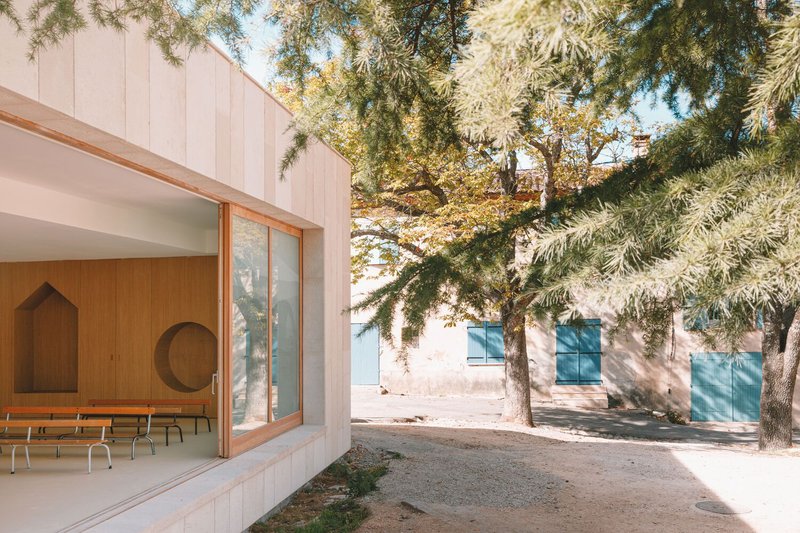 Glass doors opening from an interior wood-lined space to a gravel courtyard shaded by mature pines