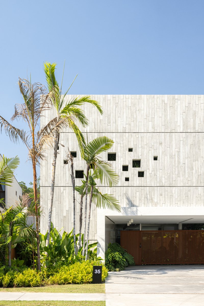 Board-formed concrete facade with scattered square windows and timber garage doors behind palm trees
