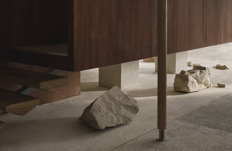 Detail of brass-trimmed walnut panel base with rough stone boulder casting shadow on terrazzo