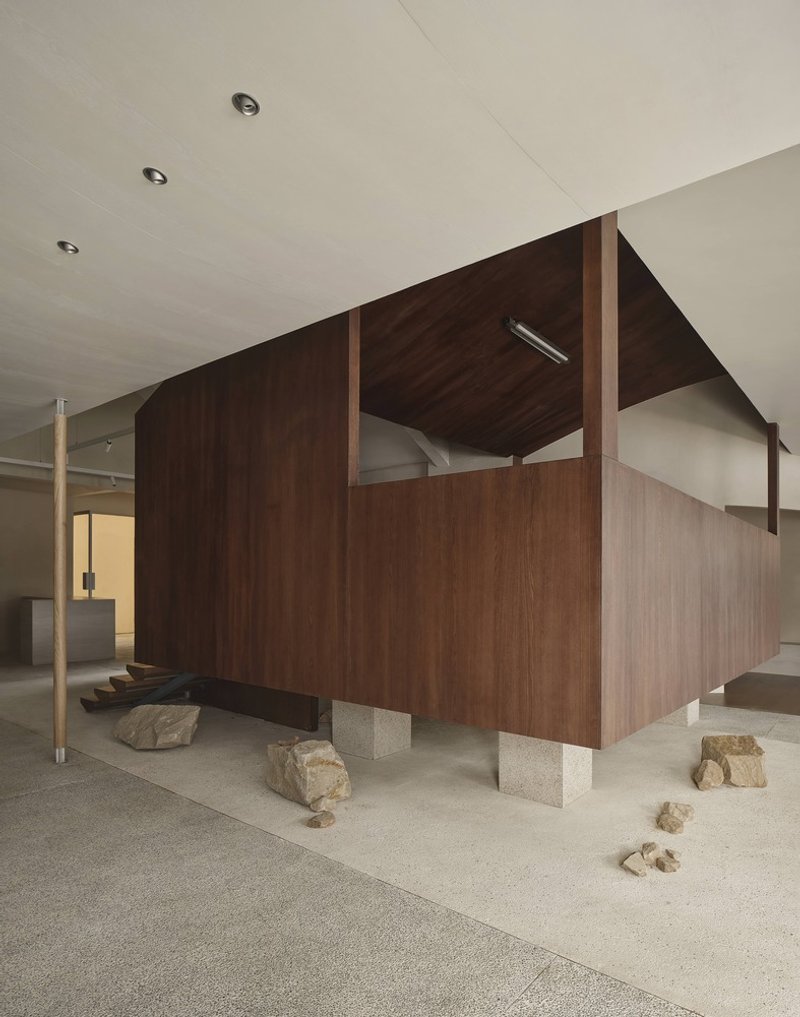 Freestanding walnut pavilion raised on concrete plinths with scattered rocks on terrazzo flooring