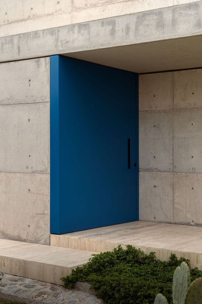 Blue pivot door set within board-formed concrete frame above planted bed with cacti and shrubs