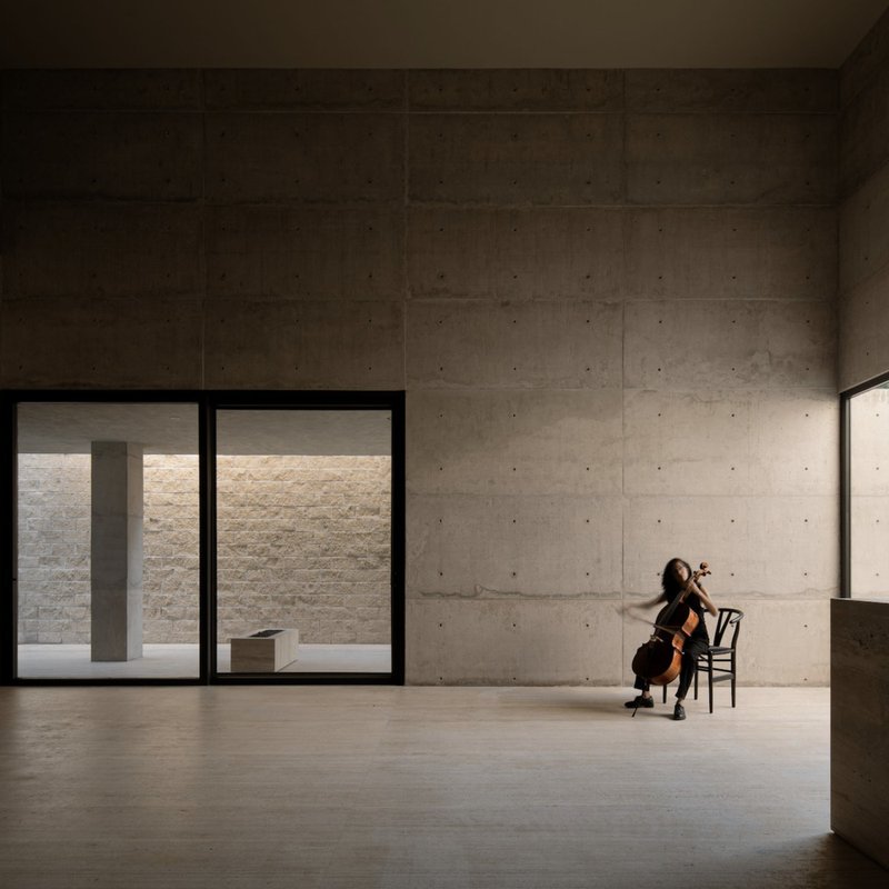 Board-formed concrete interior with cellist seated by floor-to-ceiling glazing overlooking a courtyard