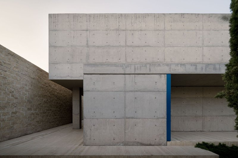 Entrance courtyard framed by board-formed concrete walls with a recessed blue door under overcast light