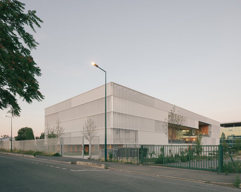 Street view of the louvered white volume behind metal fencing and a lit streetlamp at dusk