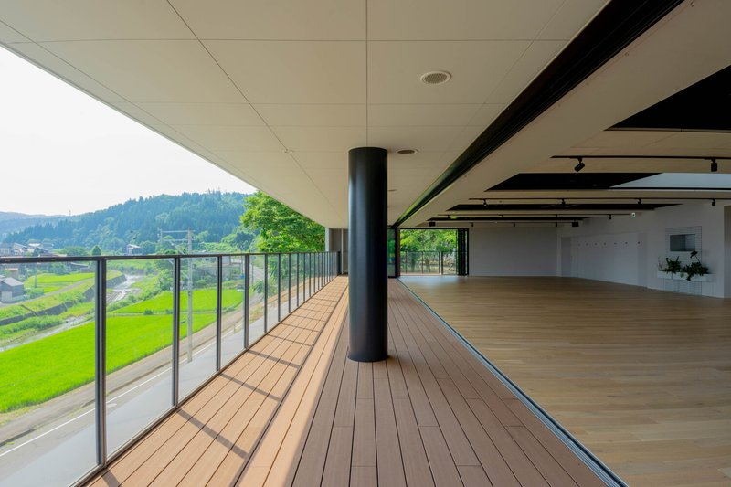 Covered terrace with timber deck and black cylindrical column overlooking green rice paddies and distant hills