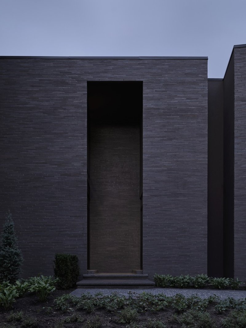Dark brick facade with recessed timber entrance door flanked by low evergreen plantings at dusk