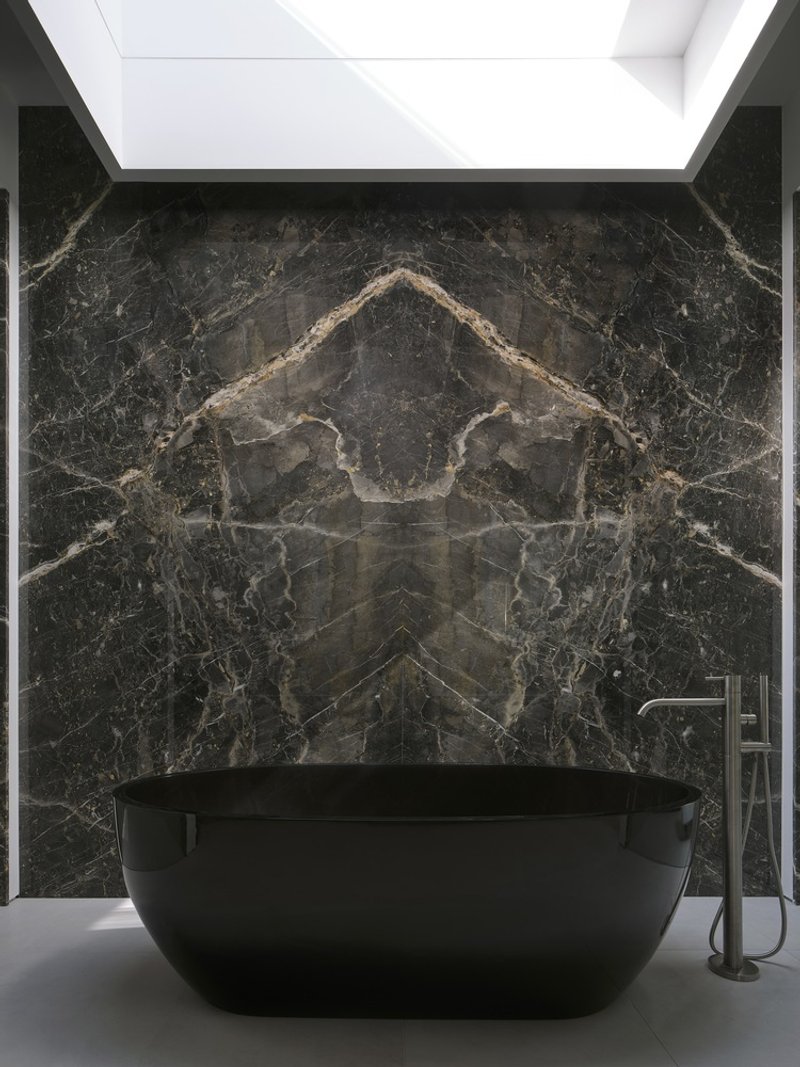Freestanding black bathtub against a bookmatched dark marble wall beneath a recessed skylight