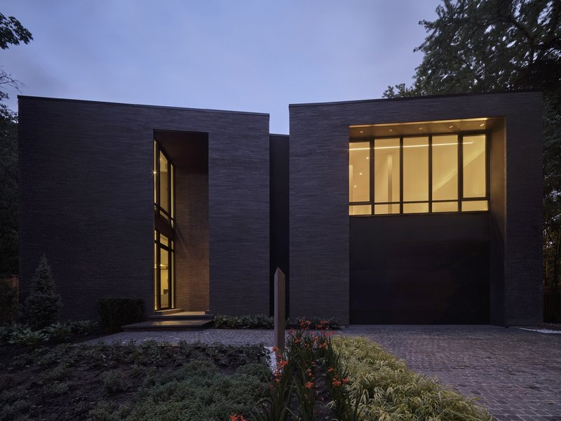 Dark brick facade with illuminated interior rooms visible at dusk beside planted beds