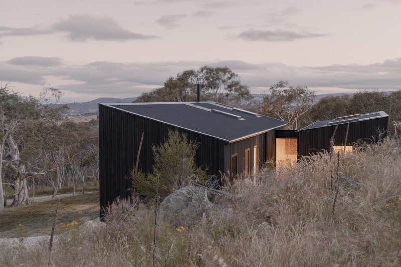Black corrugated metal volumes nestled into a hillside with native grasses under an overcast dusk sky