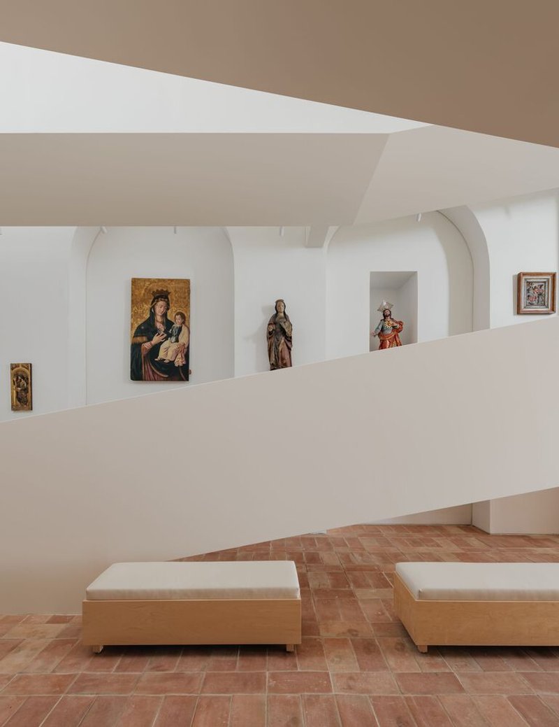 Gallery wall with arched niches displaying religious artworks above upholstered benches on terracotta tile flooring