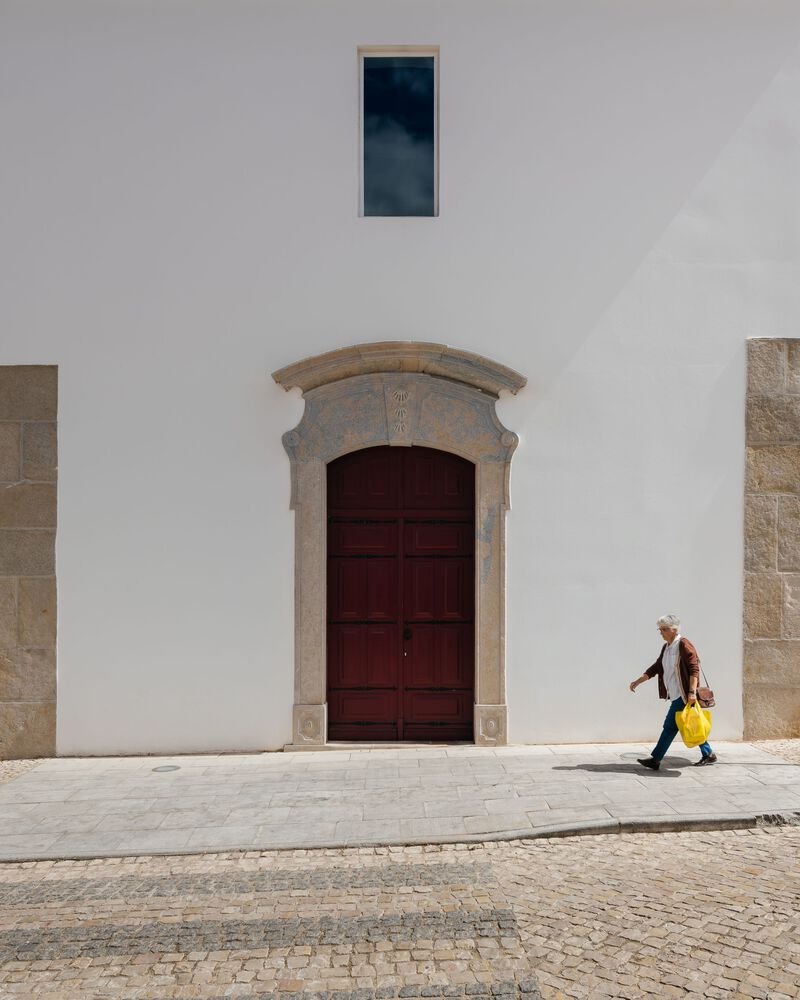 White stucco facade with stone-framed wooden door and narrow window as a pedestrian passes