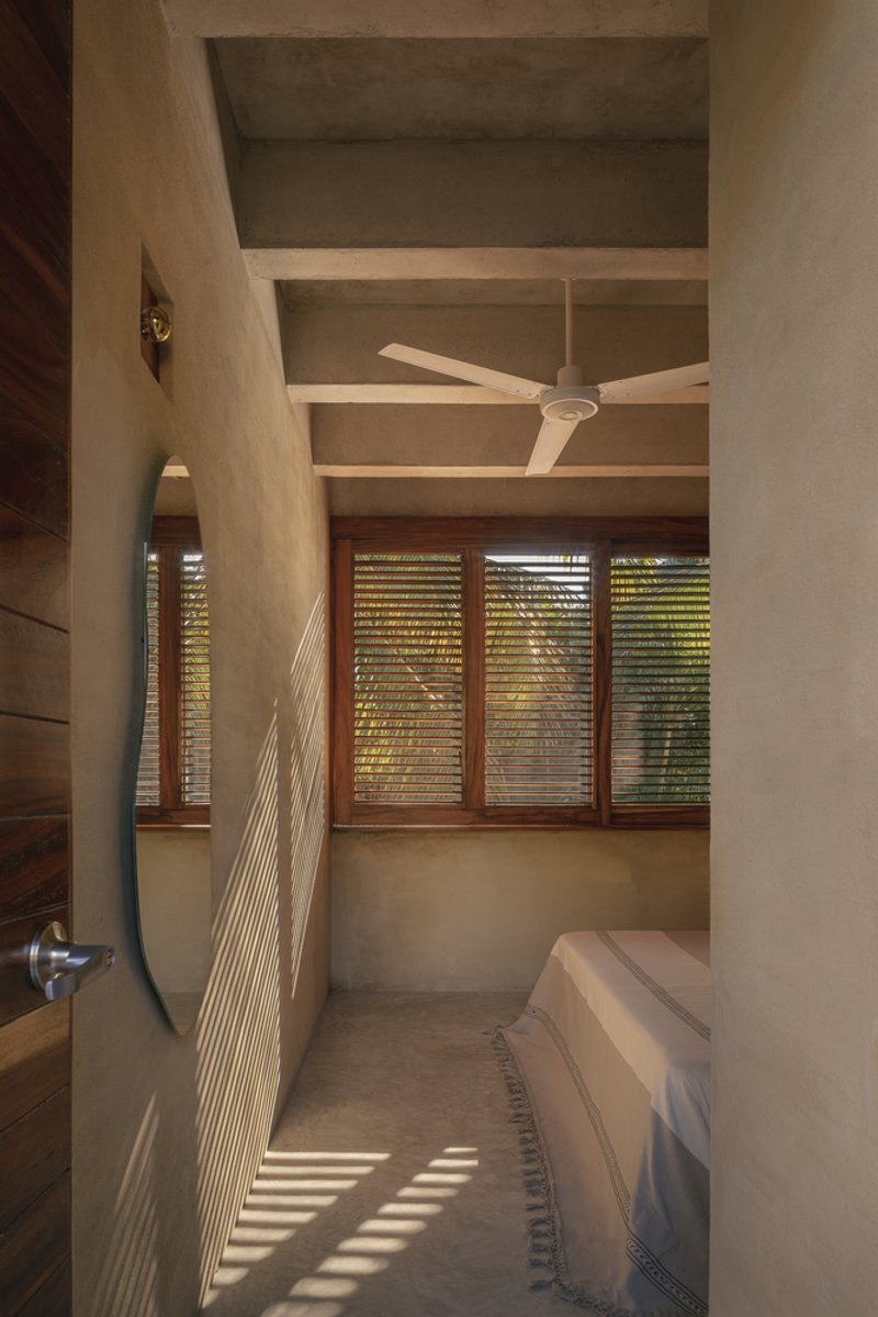 Bedroom with exposed concrete beam ceiling and timber louvered windows filtering afternoon sunlight across floor