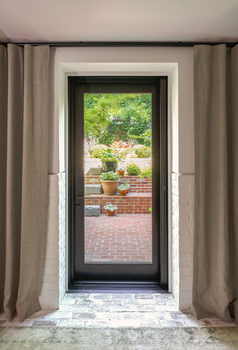 Black-framed glass door with linen curtains opening onto a brick courtyard with potted plants