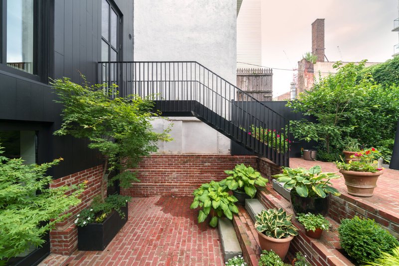 Terraced brick courtyard with planted beds and black metal exterior stair beneath lush greenery