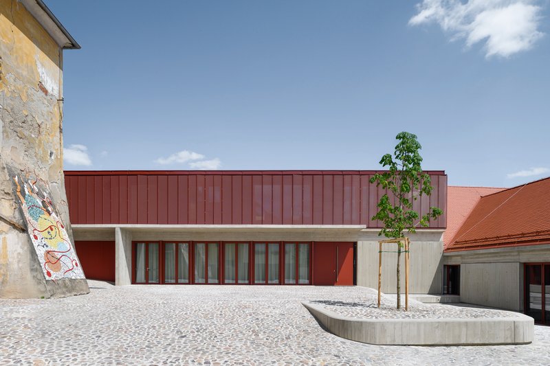 Courtyard view with red metal-clad facade and curved concrete planter holding a young tree
